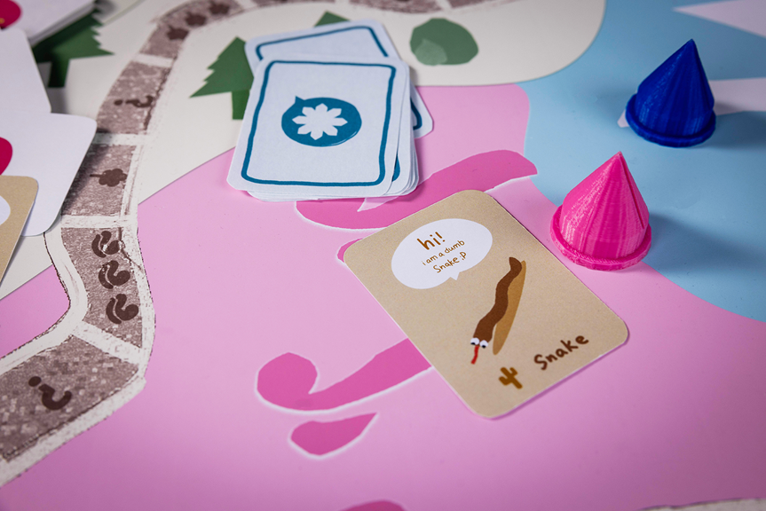 A close-up photo of a colourful board game. There is a deck of cards turned face down, pink and blue game pieces, and one card face up that shows a drawing of a snake.