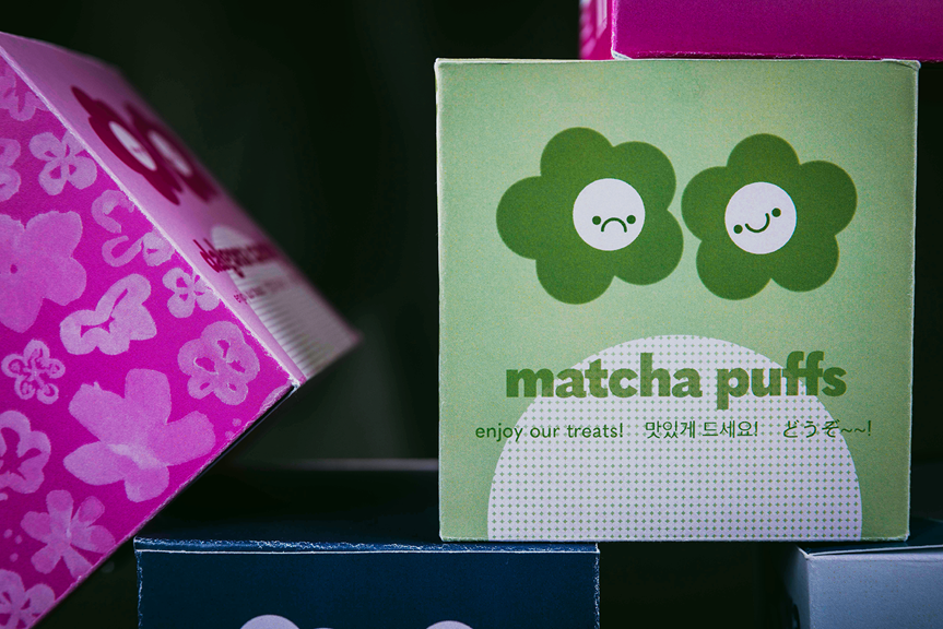 A close-up photograph of small coloured boxes of packaging. The image focuses primarily on a green box, which has text reading ‘Matcha Puffs’ and two cartoon flowers.