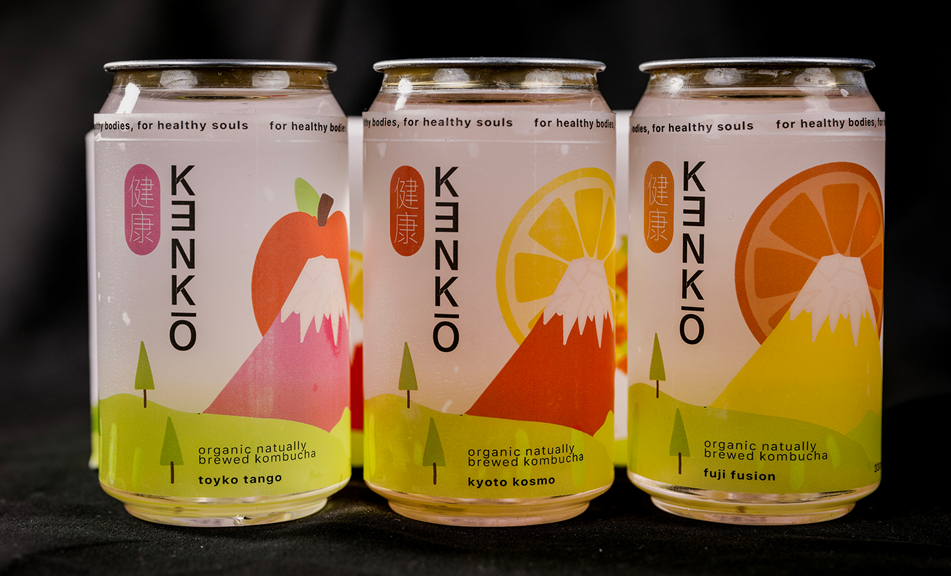 A close-up photograph of three drink cans, all labelled with KENKŌ branding. Each can has a different coloured label to represent a different flavour of kombucha.