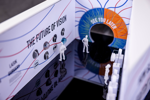 A close-up photo of an architectural model, depicting people in a gallery space. The wall reads ‘The Future of Vision’.