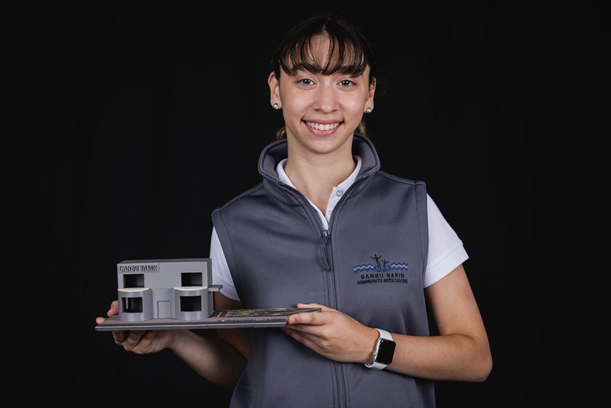 A photo of a person smiling and wearing a grey vest that has a Ganbu Barin logo. They hold a 3D architectural model of the Ganbu Barin Community Arts Centre.