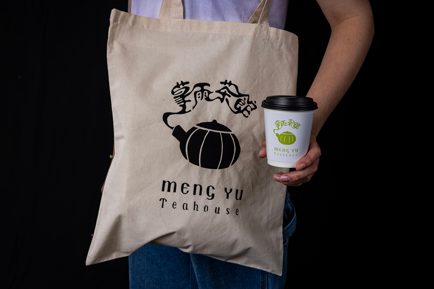 A person with a branded Meng Yu Teahouse tote bag over their shoulder. They hold a takeaway cup with a green Meng Yu Teahouse logo on it.