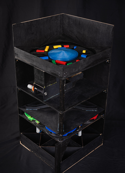 The photo shows a tall, rectangular black system with four layers. Red, blue, yellow, and green LEGO pieces are sitting on top.