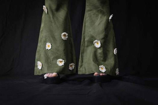 A close up photograph of the final product, showing the bottom of wide trouser legs. The trousers are olive green and have knitted daisies attached.