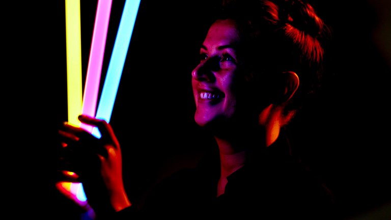 Person holds three illuminated glow sticks in a variety of colours near their face. 