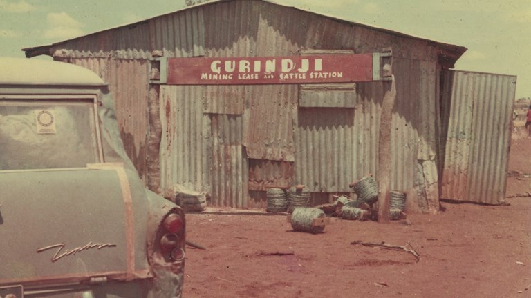 Rusty iron shed bearing a sign reading 'Gurindji Mining Lease and Cattle Station'. A Ford Zephyr is parked out the front.