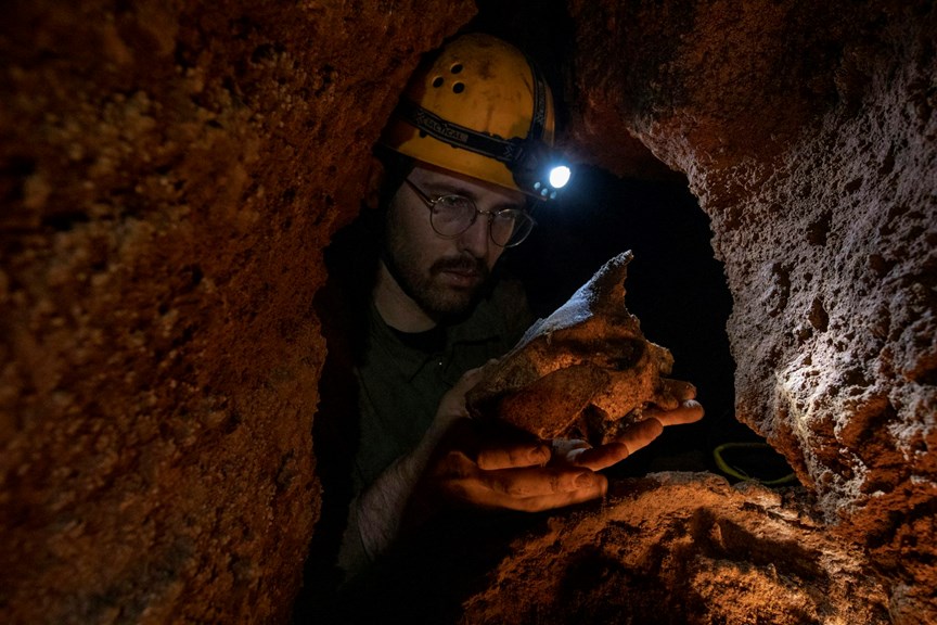 A man in a dark cave wearing glasses and a hard hat with a light on it inspects a large bone