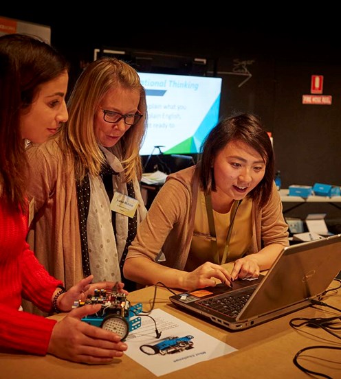 Two female teachers look on as a female museum staff member uses the mouse pad on a laptop. One teacher looks down at a blue car shaped robot with electronics on top.