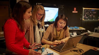 Two female teachers look on as a female museum staff member uses the mouse pad on a laptop. One teacher looks down at a blue car shaped robot with electronics on top.