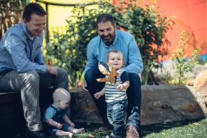 Two men and a baby, and a toddler with a leaf, sitting in the garden