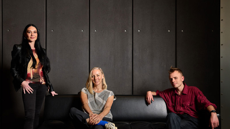One person standing next to a black couch and two people sitting on the couch in front of a black wall.  