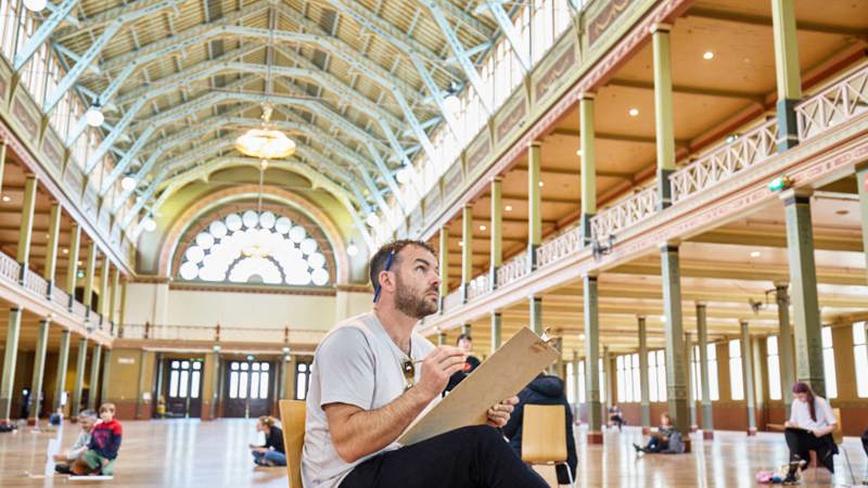 A man with a pen behind his ear sits on a chair sketching the ornate features of the Royal Exhibition Building interior.
