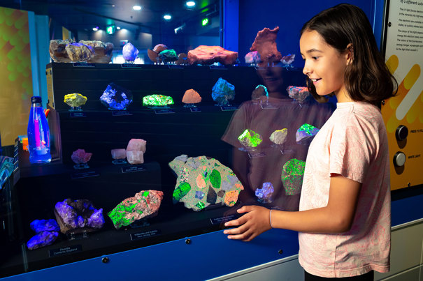 A child stands in front of a museum display case filled with fluorescent minerals glowing in bright colours under ultraviolet light. The rocks are arranged on multiple shelves, illuminating shades of green, blue, purple, and orange. The child looks closely at the exhibit, with reflections of the glowing minerals visible on the glass. 