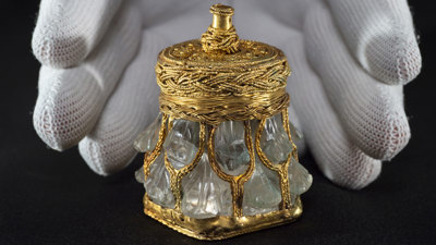 A close-up image of a hand, wearing a white glove, holding a rock crystal jar. This rock crystal jar is one of the treasures of the Galloway Hoard. It is a small, delicate item carved from crystal and decorated with gold.  