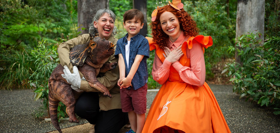 Emma Memma in an orange dress besides a performer with a baby triceratops and a child in the forest gallery.
