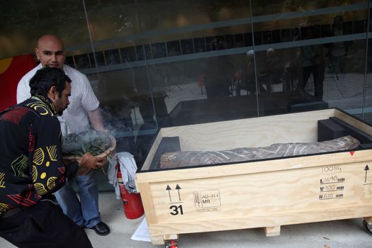 Two men with a bowl of smoking eucalyptus leaves near a box containing part of a carved tree