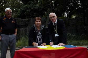 A woman and a man signing papers at a table draped in the Aboriginal flag. Another man looks on smiling.