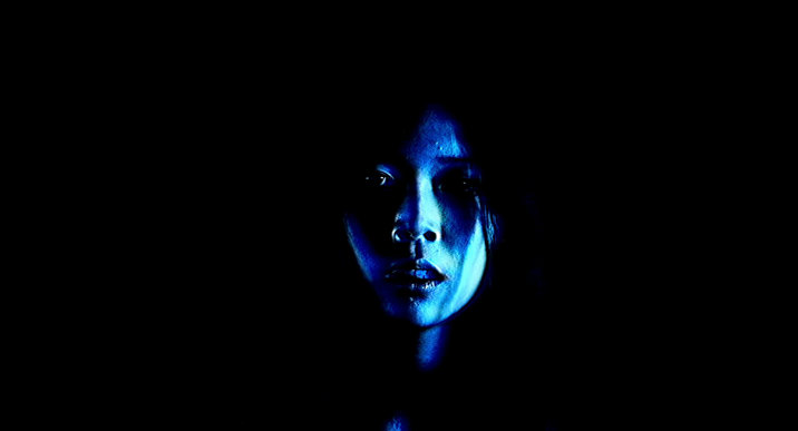 Film still showing a face illuminated by dramatic blue lighting against a black background, creating strong shadows and contrast.