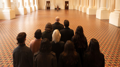 Group of performers viewed from behind, facing a lone figure across a large historic room.
