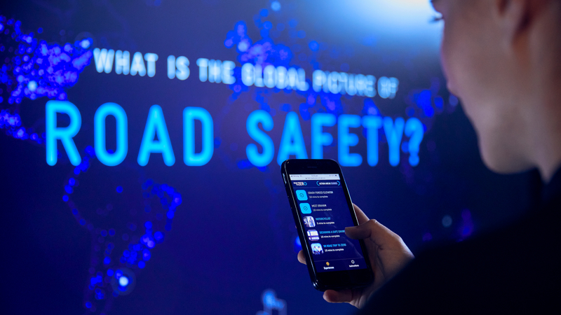 A student in the Road to Zero Experience Space holding a mobile phone standing in front of a touch screen 