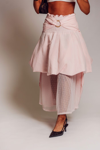 Model wearing a pale pink tiered skirt with a wrapped waistband and gold-toned ring detail, paired with black heels against a neutral backdrop.