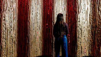 A person stands facing a tall, textured wall installation with alternating vertical panels in earthy tones of beige and deep red. The panels have organic, bark-like patterns with small glowing red lights embedded throughout, creating a dramatic and immersive effect. The floor features a mottled design in shades of gray and rust, complementing the natural theme of the display.