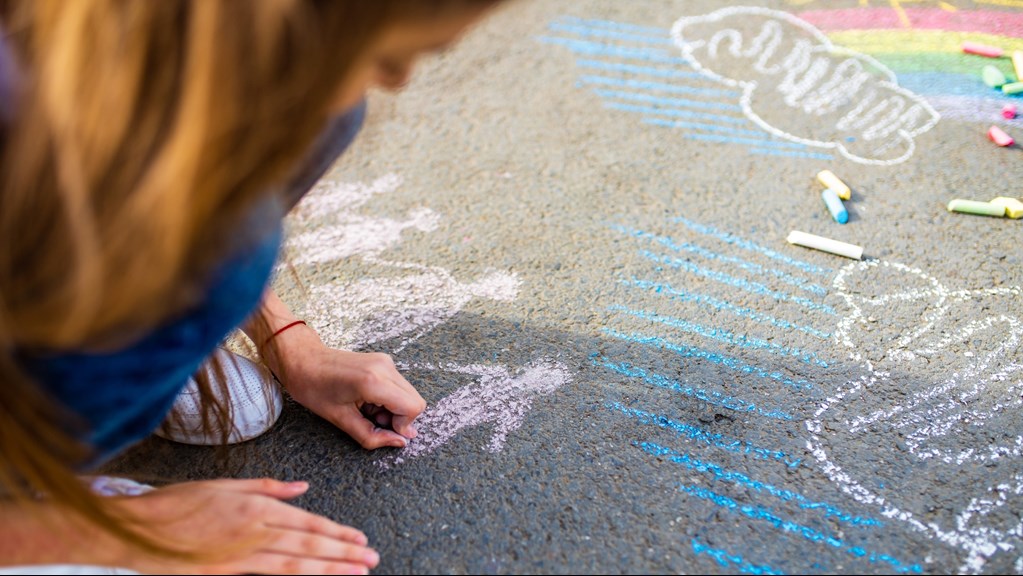 Crouching girl holds chalk in hand and draws human figures on concrete surface. 