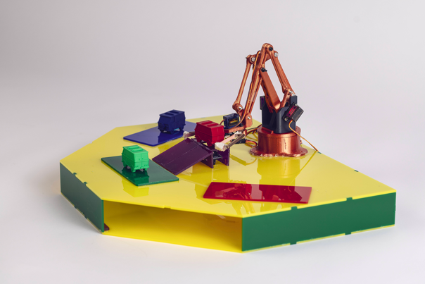 Small robotic arm on a yellow hexagonal platform, sorting model trucks in a variety of colours.