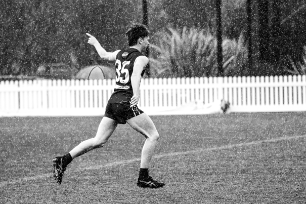 Black and white photo of young man in AFL uniform, running across a football field in the rain as he raises an index finger to the sky.