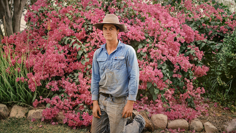 A man in gardening clothes and sun hat kneels in front of a pink-blossoming bush.