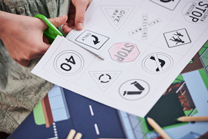 A child’s hands using scissors to cut out mini paper road signs that they have coloured in.