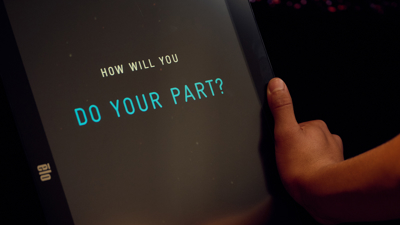 A touchscreen in the Road to Zero Experience Space with the text How Will You Do Your Part? on the screen near a student’s hand 