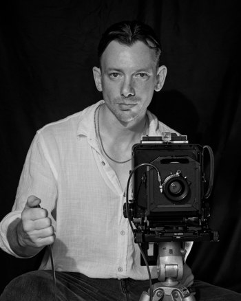 Headshot of David Charles Collins holding an old tripod mounted camera..