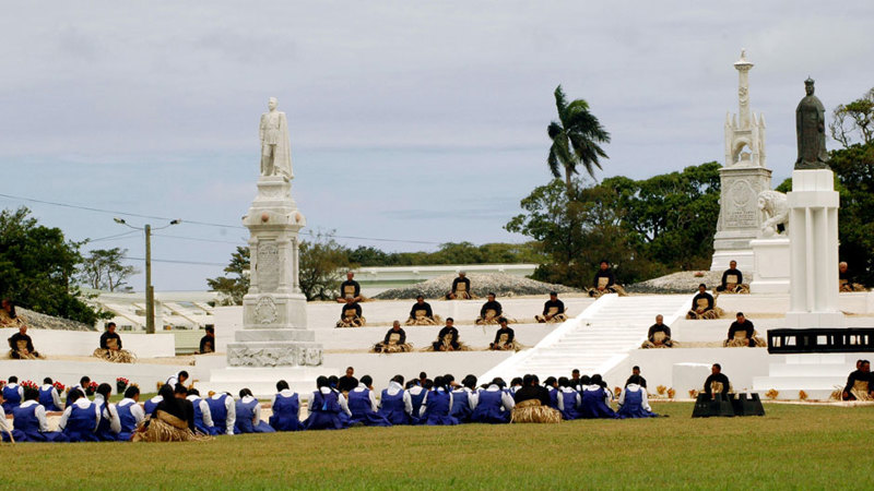 People gather at a tomb. There group dressed in black sitting on the steps of the monument, there is another group dressed in blue sitting on the grass facing the monument.