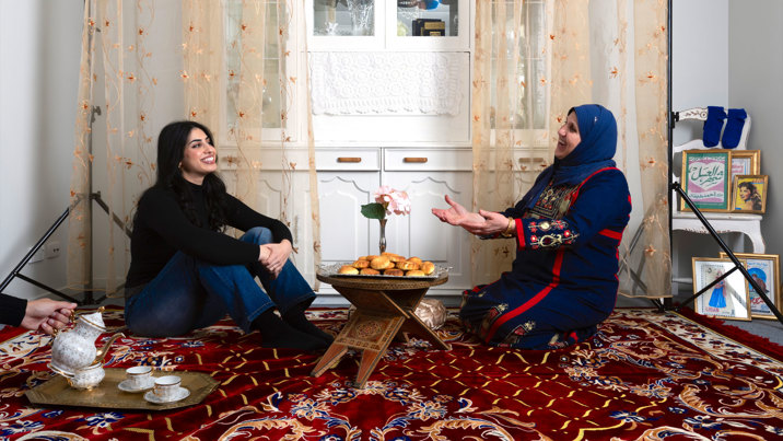 In a white walled living room, on an ornate red rug, a smiling younger women in jeans and a shirt sits across from a smiling older women wearing navy blue traditional Turkish clothing, with her arms outstretched. In between them is a bowl of bread on a wooden stand and to the side a hand pours tea into fine white China cups on as gold tray. In the background is a carved white display cupboard, translucent beige curtains and a chair with gold-framed photos of women and Arabic writing.