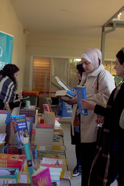 A group of women, some with hijabs and some without, browse a table of Muslim-focused literature from a book vendor.