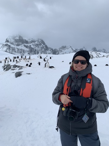 A woman in Antarctic clothing stands on ice with penguins in the background.