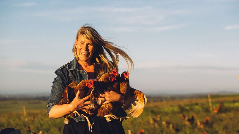 Woman standing in front of a chicken coop, holding two chickens in her arms as other chickens graze on the ground