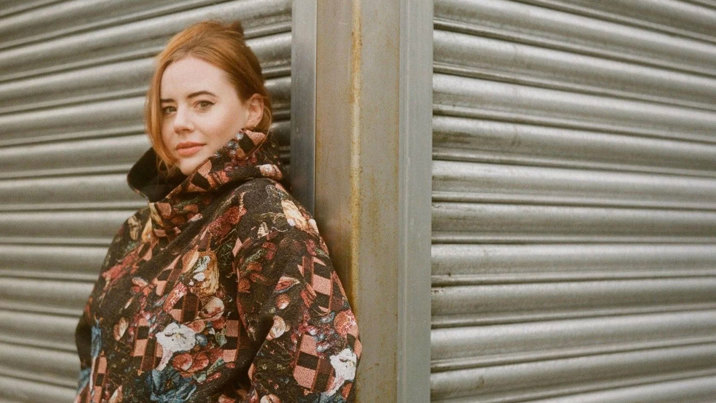 Lotte leans on a metal wall and wears a jacket with a floral pattern.