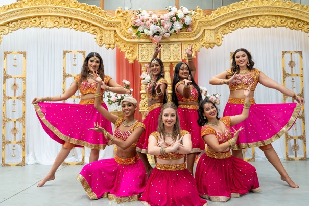 Seven Bollywood dancers posing for a photo wearing pink sequined dresses and tops.