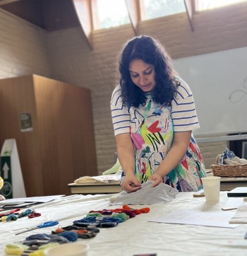 Manjari stands in front of a tables with textiles and threads and works on her embroidery. Manjari wears a dress with colourful abstract patterns.