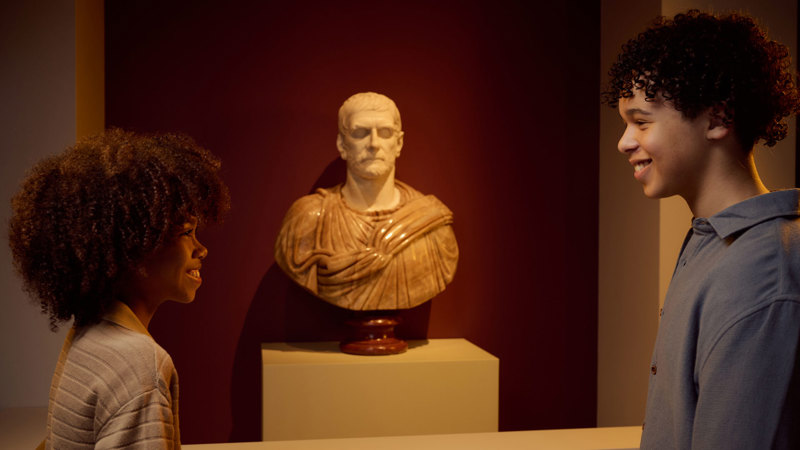 Two children smiling at each other in front of a Roman marble bust in ROME: Empire, People Power People Exhibition at Melbourne Museum