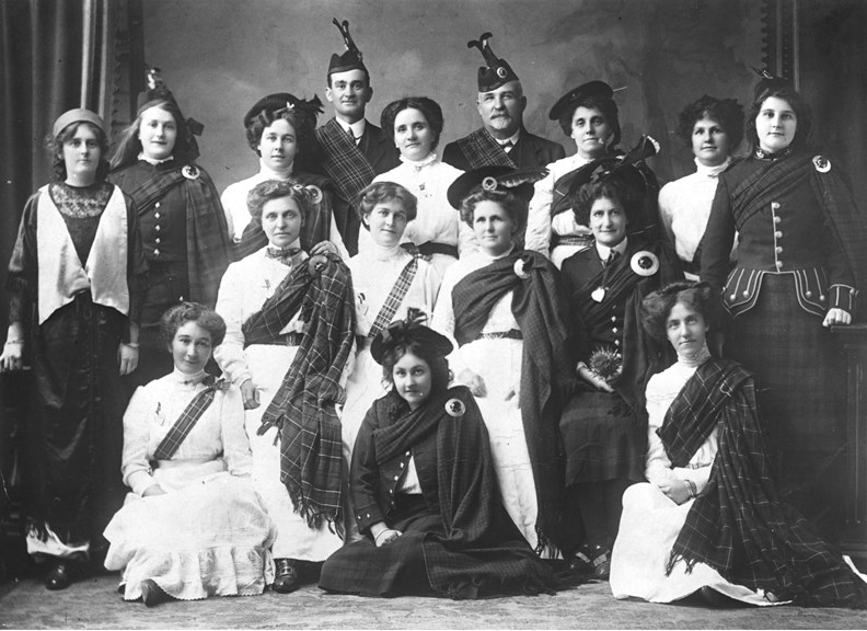 Late 19th century Victorian Caledonian Society in Bendigo. A group of women in Highland costumes. Most wear white dresses with tartan sashes while three wear long kilts. There are two men (pipers?) at the rear of the group.