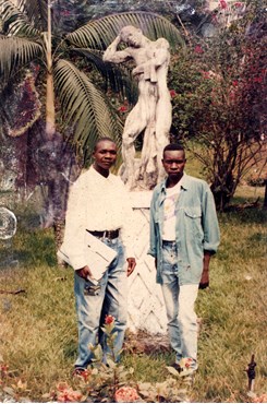 Nickel Mundabi & Mpuon Ngadwa standing in front of a figurative sculpture in the grounds of the Congo Academy. A palm and other trees are visible in the background. The man on the left is holding a folio. 