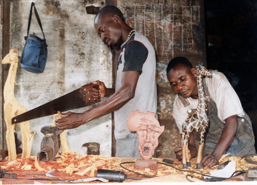 Two sculptors in a woodwork workshop. One man is standing using a saw to the left of the workbench, and the other man is working on fine detail of a carved giraffe sculpture, to the right. The workbench has an array of tools and sculptures in progress surrounded by wood shavings.