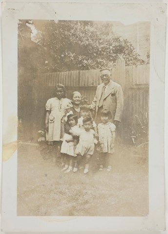 Photo of Sydney and May Gung, and five of their grandchildren taken in a back yard in front of fence.