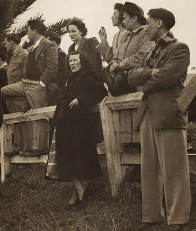 Row of nine people  (two in front of fence, the rest behind) all watching in the same direction in an outdoor scene. 