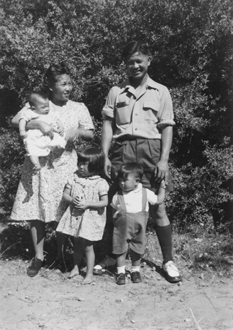 Group of two adults and three children in an outdoor setting, bushes in the background.