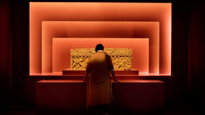 Person viewing an illuminated Roman sarcophagus framed by layered red exhibition walls. 