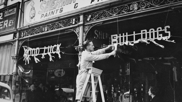 A black and white photograph of a man up a ladder outside a shop.